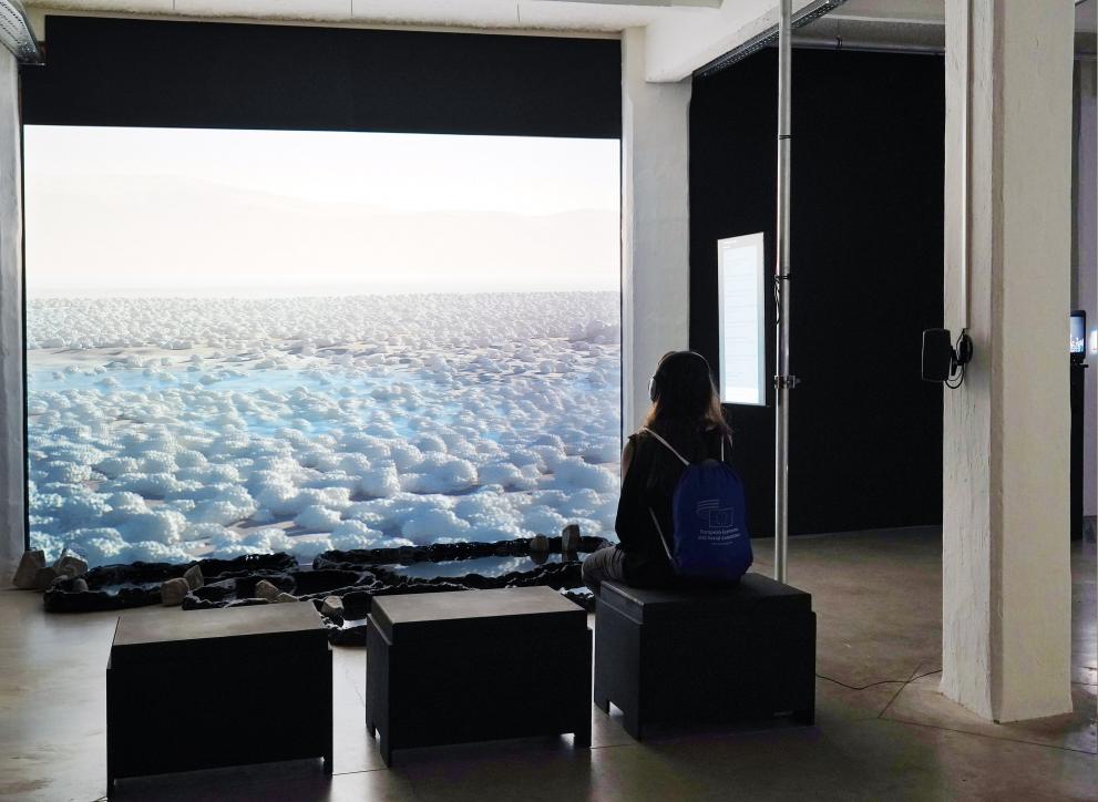 With Salt and Rocks in Our Veins being showcased at the NaturArchy exhibition. Large projected video of digital landcape of a salt saline, with person sittin on stool watching with headphones. Image credits, Penelope Cain. 